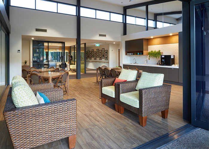 A modern communal lounge and kitchenette at Kewarra Aged Care with high ceilings, woven armchairs, and bright natural light.